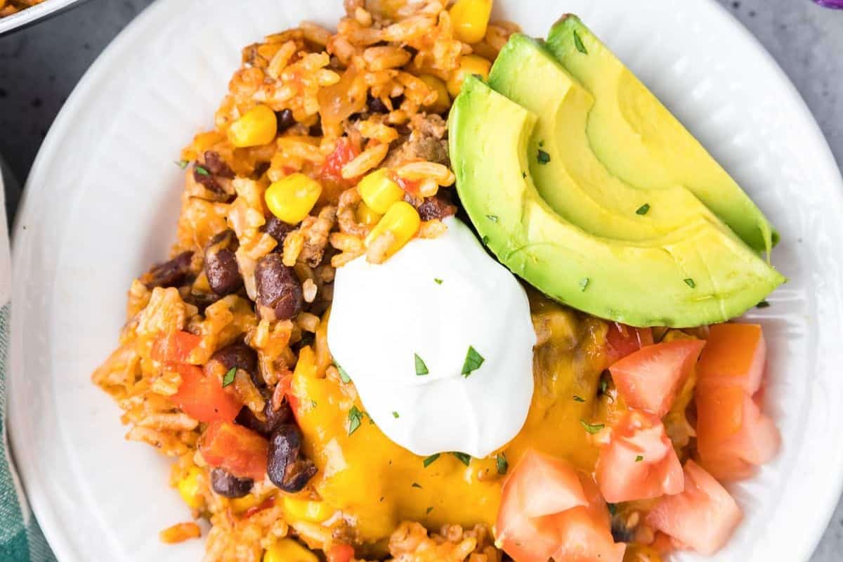 A bowl of Mexican rice with corn, black beans, diced tomatoes, melted cheese, sour cream, and sliced avocado offers an epic Cinco de Mayo delight.