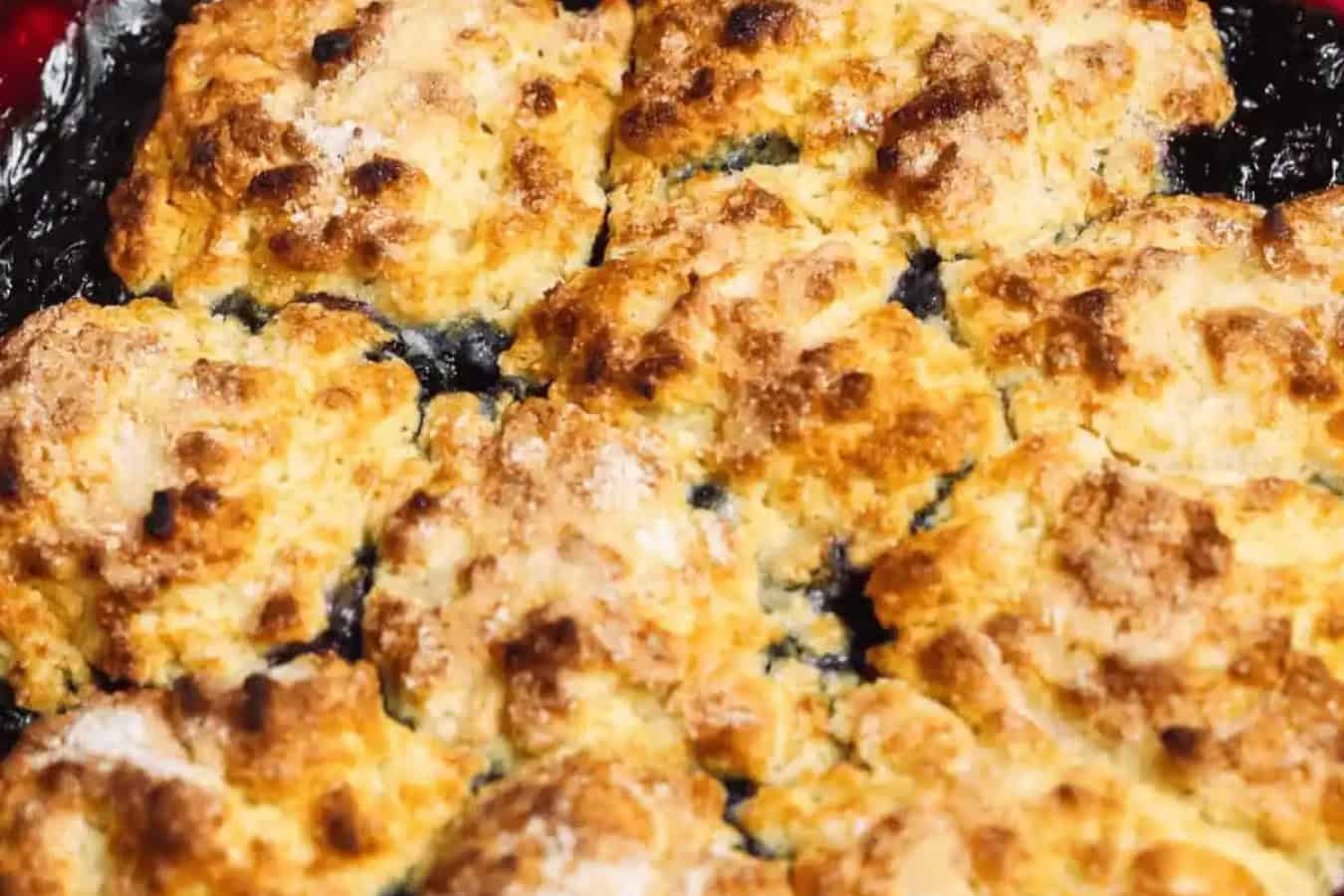 Close-up of a golden-brown, baked cobbler with a crumbling, biscuit-like topping. The dark filling beneath the topping slightly peeks through, making it one of those beautiful desserts that captures the essence of tasty indulgence.
