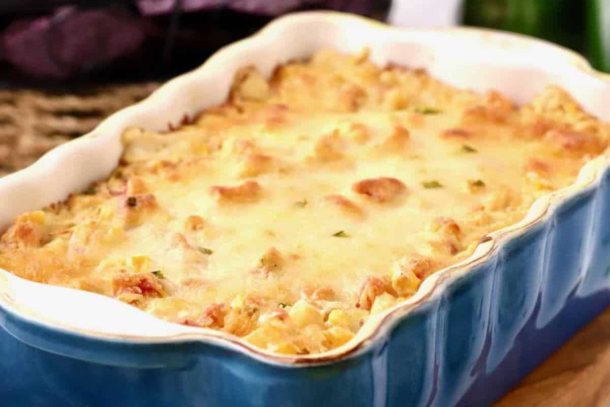 A hot corn dip in a rectangular blue dish, perfect for a Cinco de Mayo party, topped with melted cheese and small herbs, set on a woven surface.