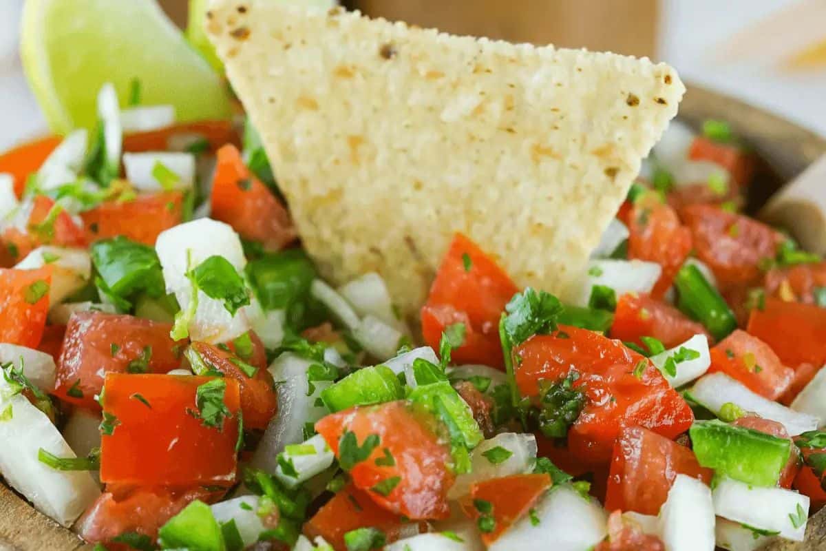 A bowl of fresh salsa with diced tomatoes, onions, and green peppers, garnished with cilantro—a perfect dip for any party. A tortilla chip is plunged into the salsa, with a lime wedge in the background, capturing a festive Cinco de Mayo vibe.