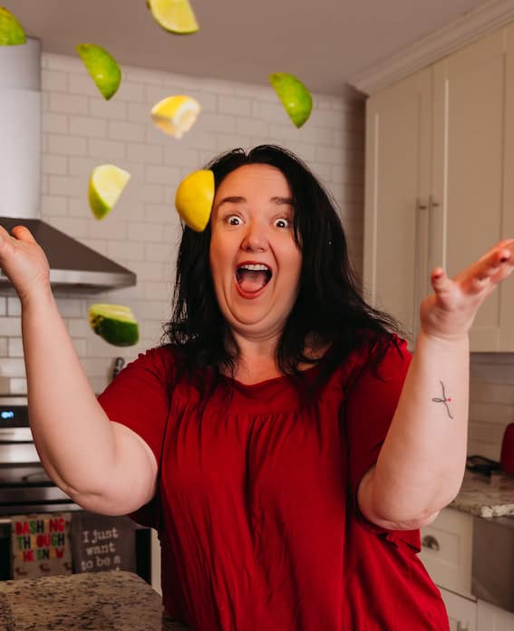 In a lively kitchen scene, a woman in a red shirt is playfully tossing lime and lemon slices as she prepares the perfect pork dish.