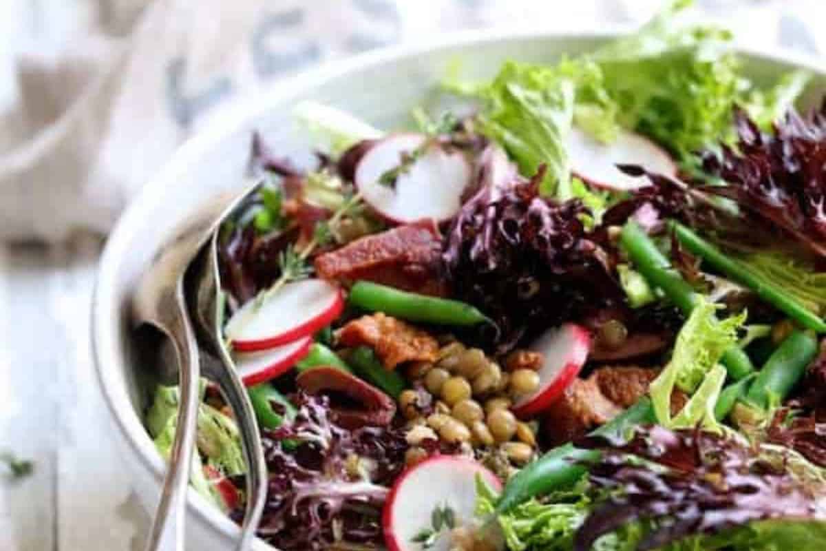 A bowl of salad with mixed greens, radish slices, lentils, bacon, and green beans embodies one of the best spring salads. Complete with a serving spoon and fork, it's an ideal choice for seasonal salad recipes.