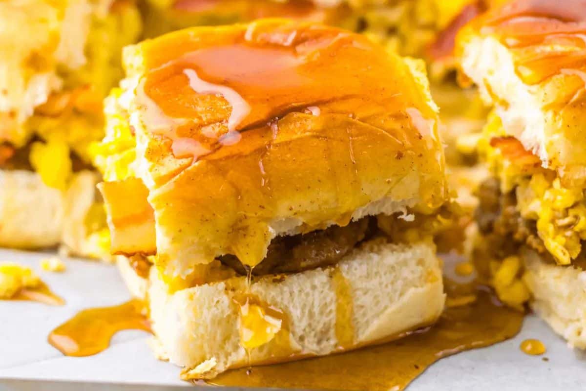 This breakfast slider, perfect for spring brunch recipes, features scrambled eggs, sausage, and bacon on a bun, topped with melted butter and drizzled with syrup.