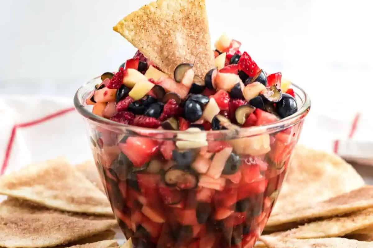 Glass bowl of mixed fruit salsa with diced apples, strawberries, blueberries, and olives. Perfect for a Cinco de Mayo fiesta, it pairs beautifully with a cinnamon sugar tortilla chip dipped in the salsa, surrounded by more chips on the plate for an irresistible dessert delight.