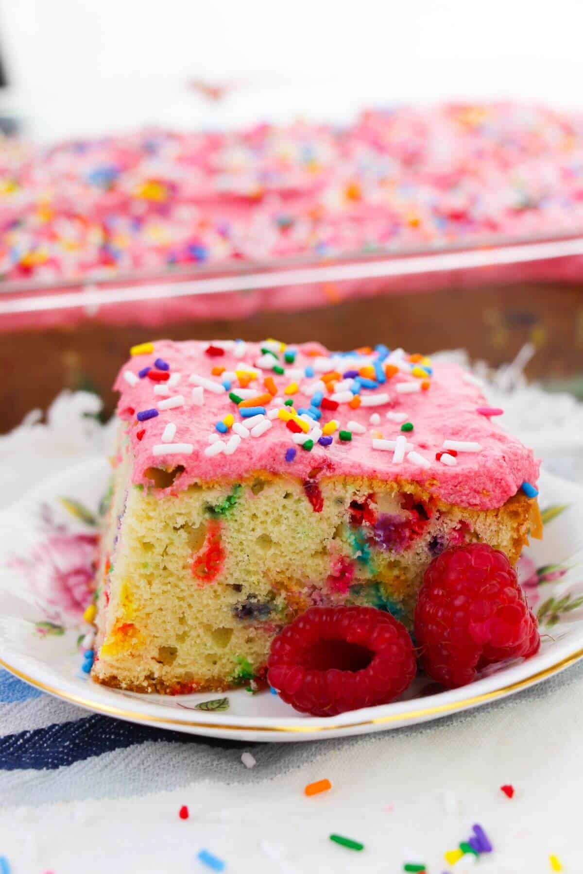 A slice  on a floral plate with two raspberries in front and a larger cake in the background.