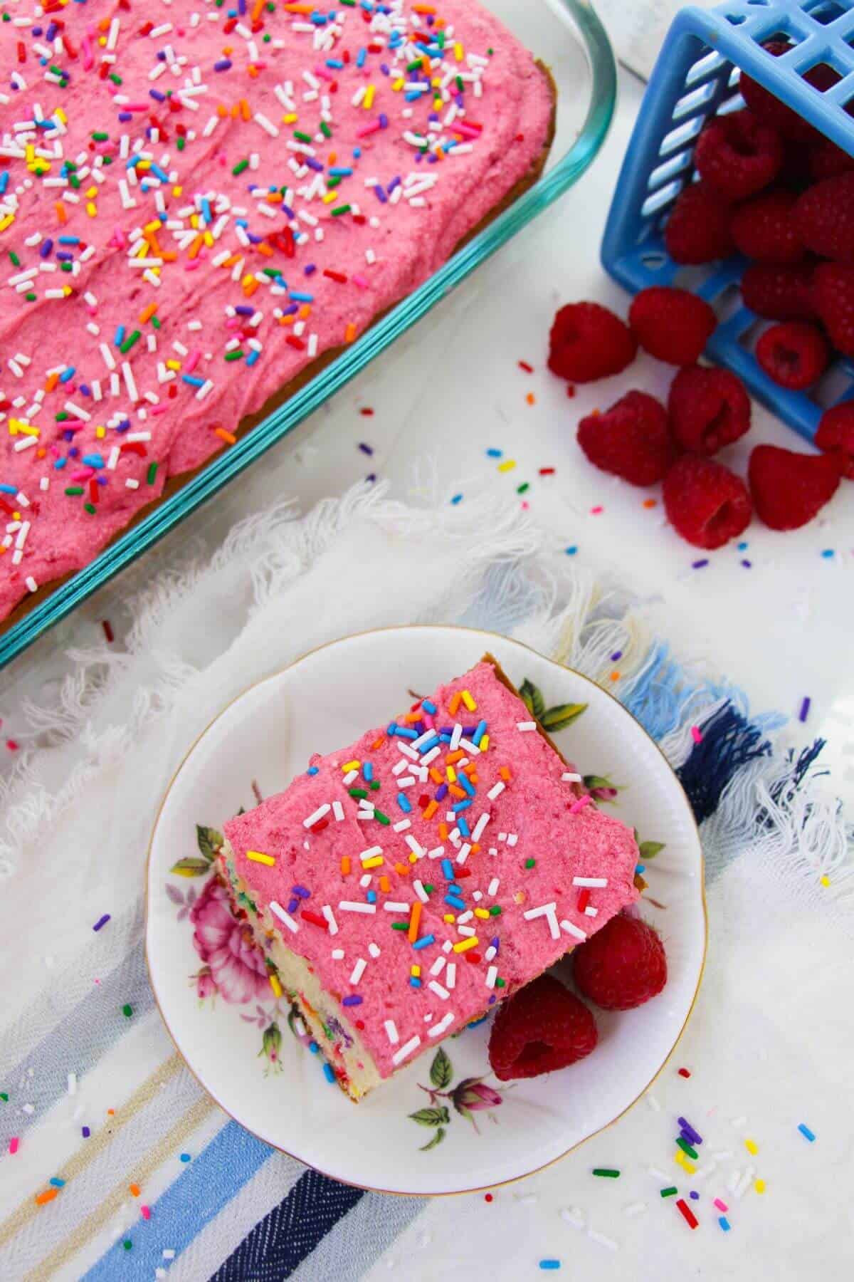 A slice  with pink frosting and sprinkles sits on a plate with raspberries, next to a baking dish of the same colorful cake and a container of fresh raspberries.