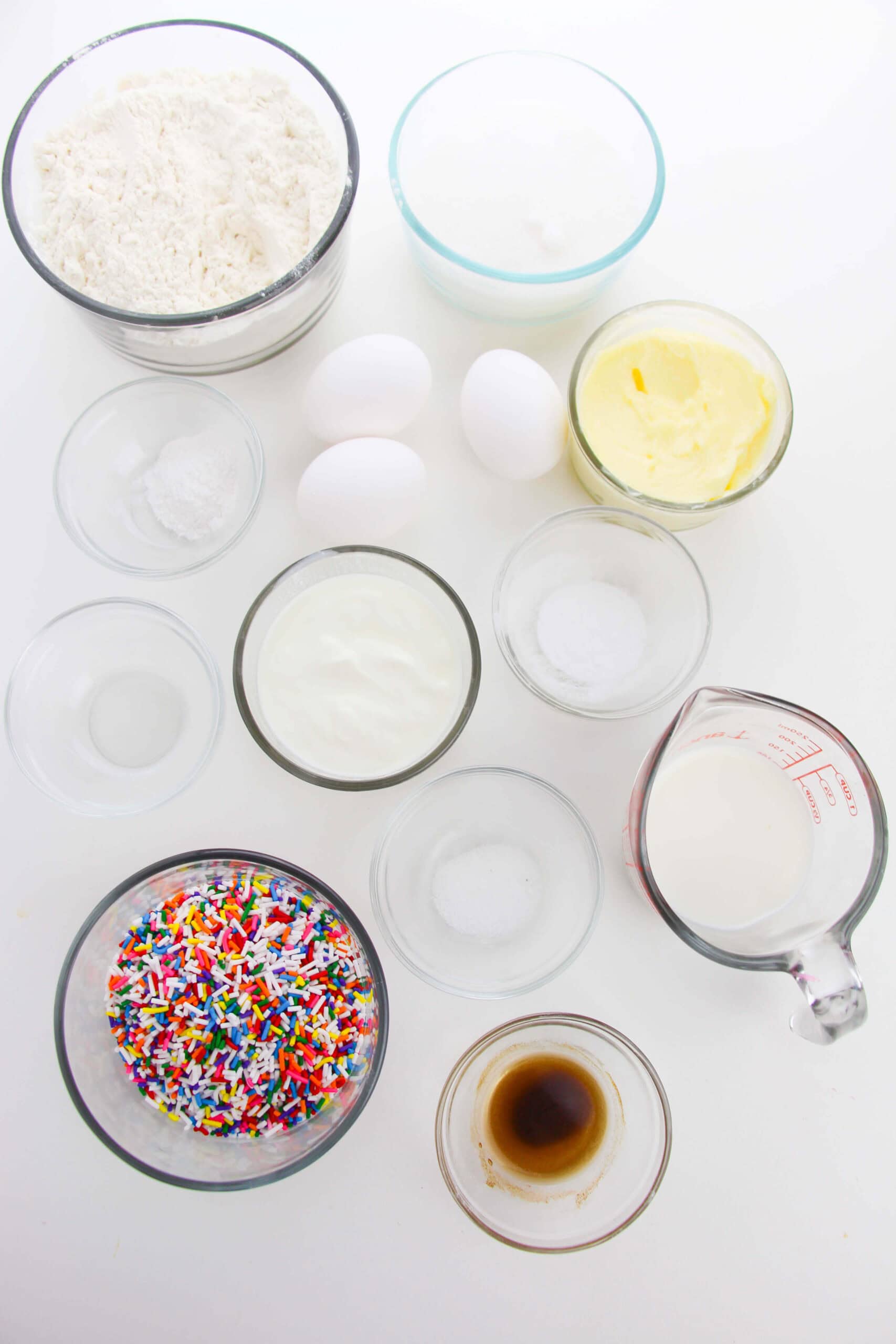 Assorted baking ingredients for Confetti Cake in clear bowls, including flour, butter, sugar, eggs, milk, vanilla extract, yogurt, baking powder, salt, and colorful sprinkles, all arranged on a white surface.