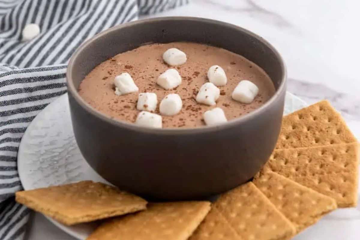 A bowl of hot chocolate dip topped with mini marshmallows, a fiesta of flavors, surrounded by graham crackers on a white plate with a striped cloth nearby.