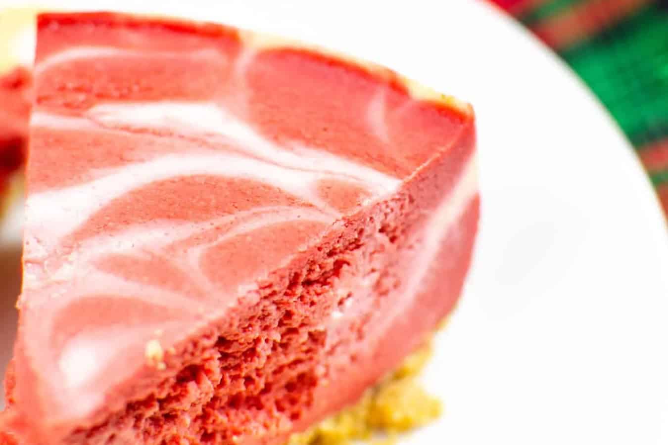 A close-up of a slice of red and white swirled cheesecake on a white plate, showcasing a textured crumbly base. This tasty dessert is a true work of art, perfect for those who appreciate beautiful desserts.