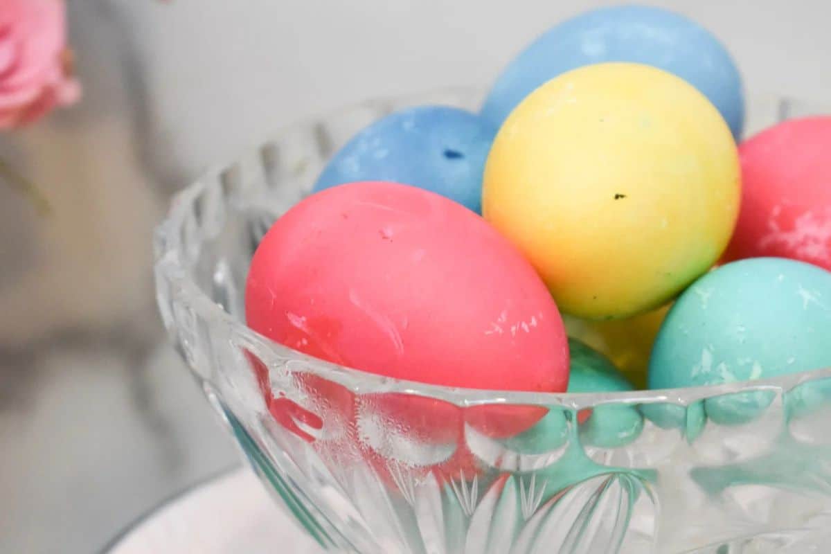 A glass bowl showcases colorful dyed eggs in vibrant shades of pink, blue, yellow, and green on a light background. Perfect for Easter 2023, this creative Easter idea adds a festive touch to your celebrations.