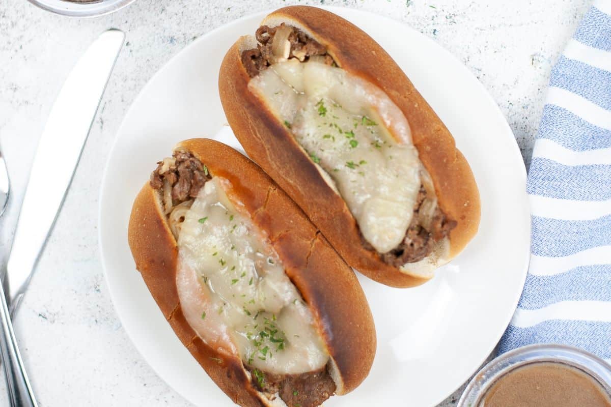 French Dip sandwiches with sliced beef and melted cheese on a white plate, with bowls and cutlery in the background.