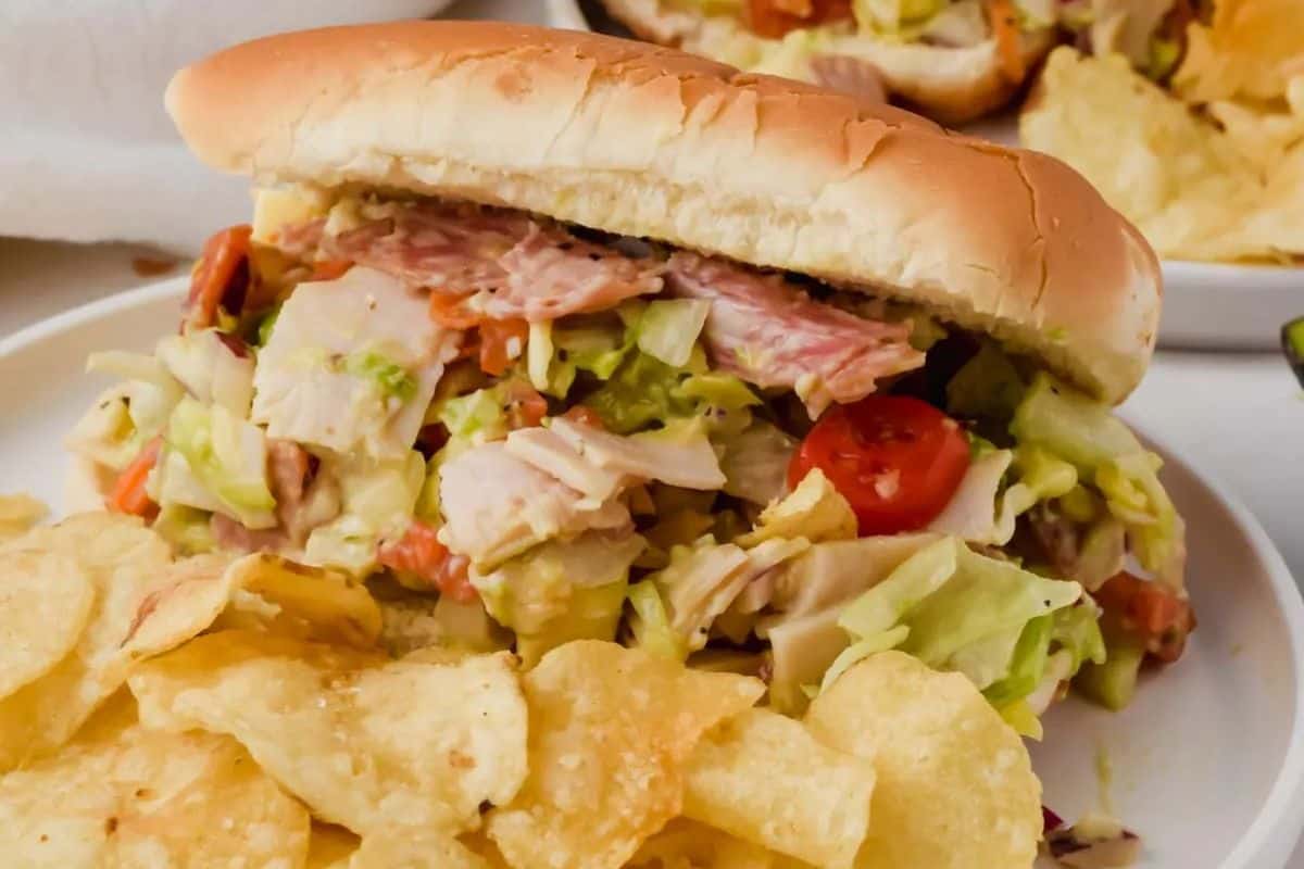 Italian grinder sandwich with sliced meats, lettuce, tomatoes, and other fillings on a long roll, served with potato chips on a white plate.