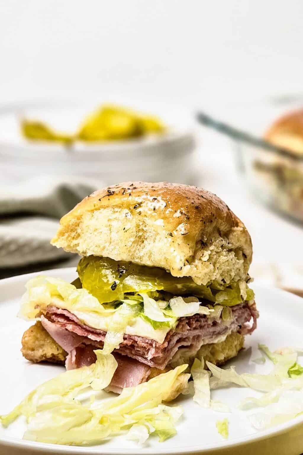 An enticing Italian slider featuring layers of meat, pickles, and crisp lettuce nestled between a fluffy bun on a plate. In the background, a bowl brims with extra pickles for an added zest.