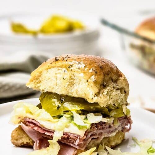 An enticing Italian slider featuring layers of meat, pickles, and crisp lettuce nestled between a fluffy bun on a plate. In the background, a bowl brims with extra pickles for an added zest.