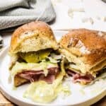 Ham and cheese sandwiches with lettuce and pickles rest on a white plate, set on a wooden surface with a napkin nearby.