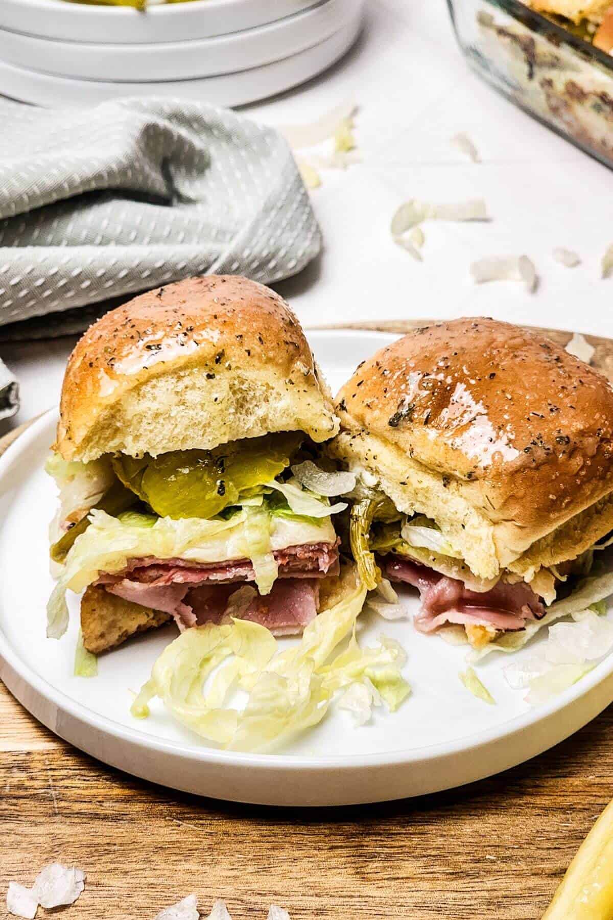 Ham and cheese sandwiches with lettuce and pickles rest on a white plate, set on a wooden surface with a napkin nearby.
