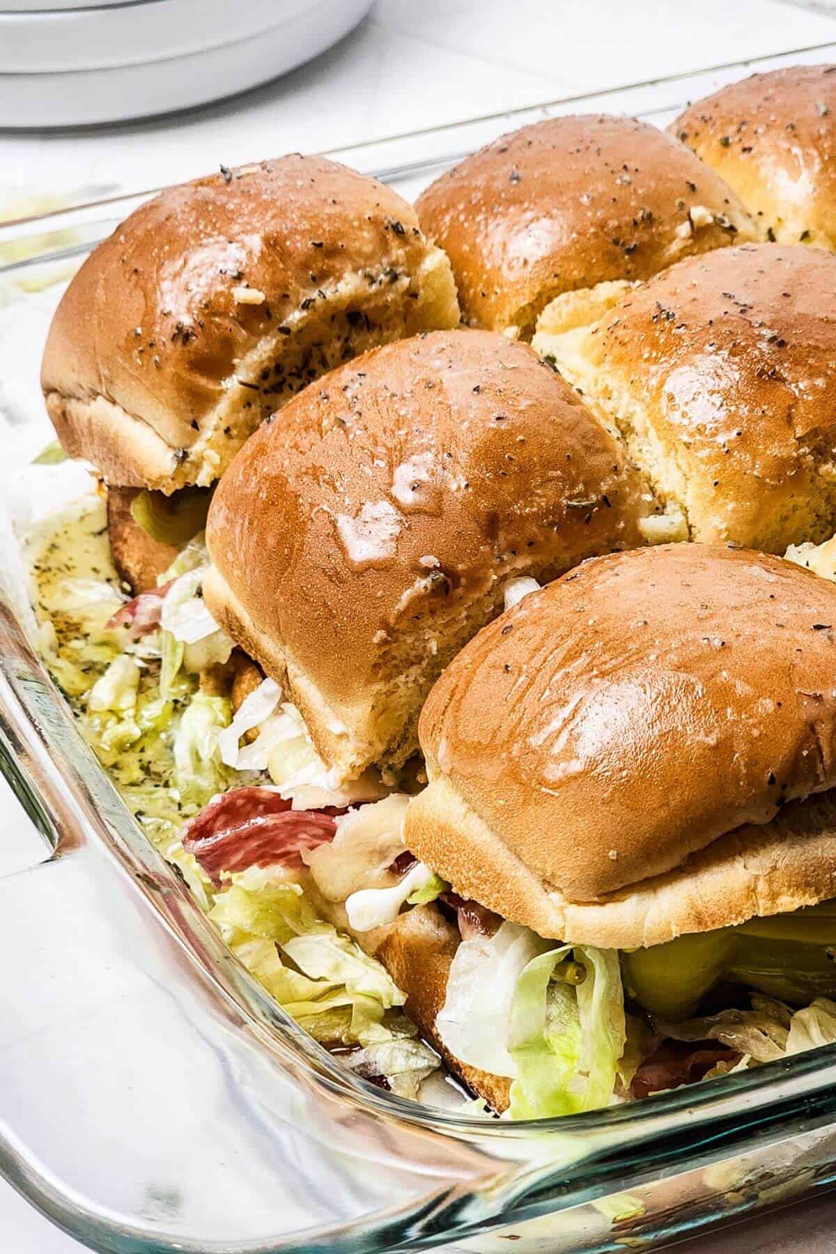 A tray of Italian sliders features baked sandwich sliders, stacked with layers of lettuce, savory meats, and pickles on soft, glazed buns.