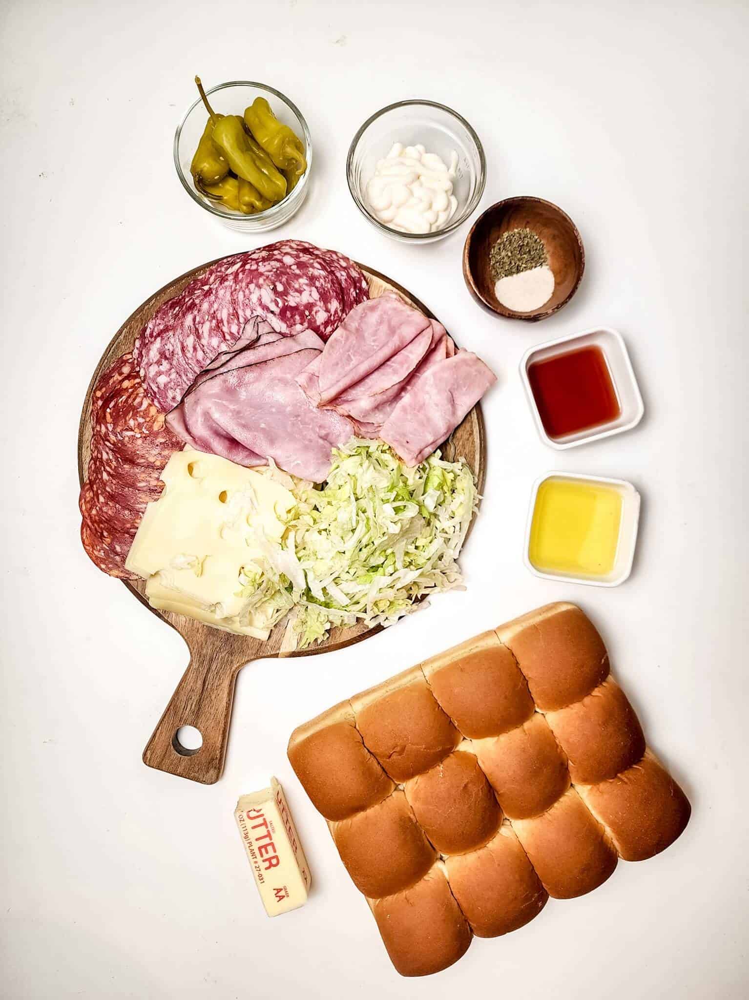 Ingredients:a wooden board with deli meats, cheese, and lettuce. Nearby are Hawaiian rolls, butter, banana peppers in a bowl, and condiments in small containers.