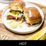 Two sizzling Italian sliders rest on a white plate with lettuce, pickles, and deli meat, all presented on a rustic wooden board. A pickle wedge is nestled nearby. At the bottom, the text proudly declares "Hot Italian Sliders.