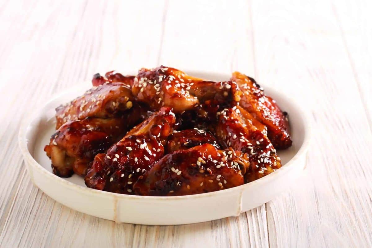 A white plate filled with delicious glazed chicken wings topped with sesame seeds rests on a light wooden surface.