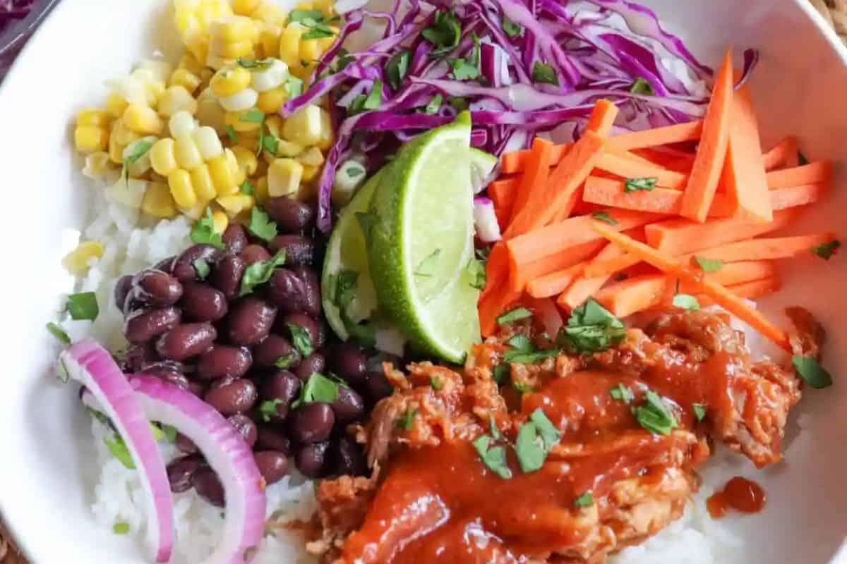 A vibrant bowl with rice topped with corn, black beans, shredded purple cabbage, carrot sticks, lime wedges, sliced red onion, and saucy shredded meat is garnished with chopped herbs—perfect for celebrating Cinco de Mayo in burrito style.