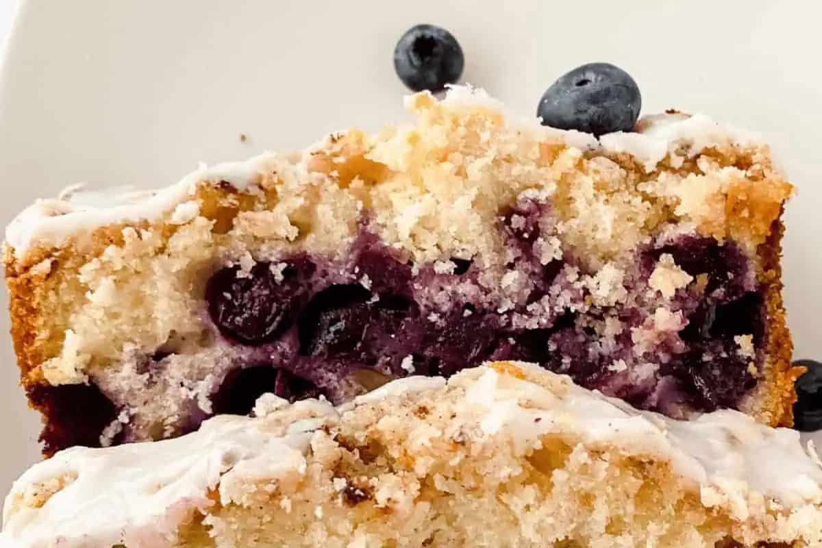 Close-up of sliced blueberry loaf with white icing, a perfect addition to spring brunch recipes. The cake features visible blueberries and crumbs, with a few fresh blueberries scattered on the plate.