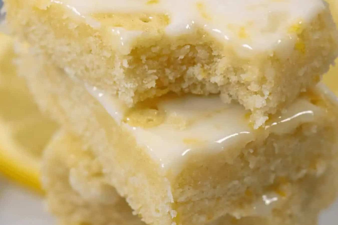 Two lemon brownies stacked, topped with a glossy lemon glaze, embody the essence of spring desserts. The top bar has a bite taken out, revealing its soft, crumbly texture—truly a beautiful dessert to savor.