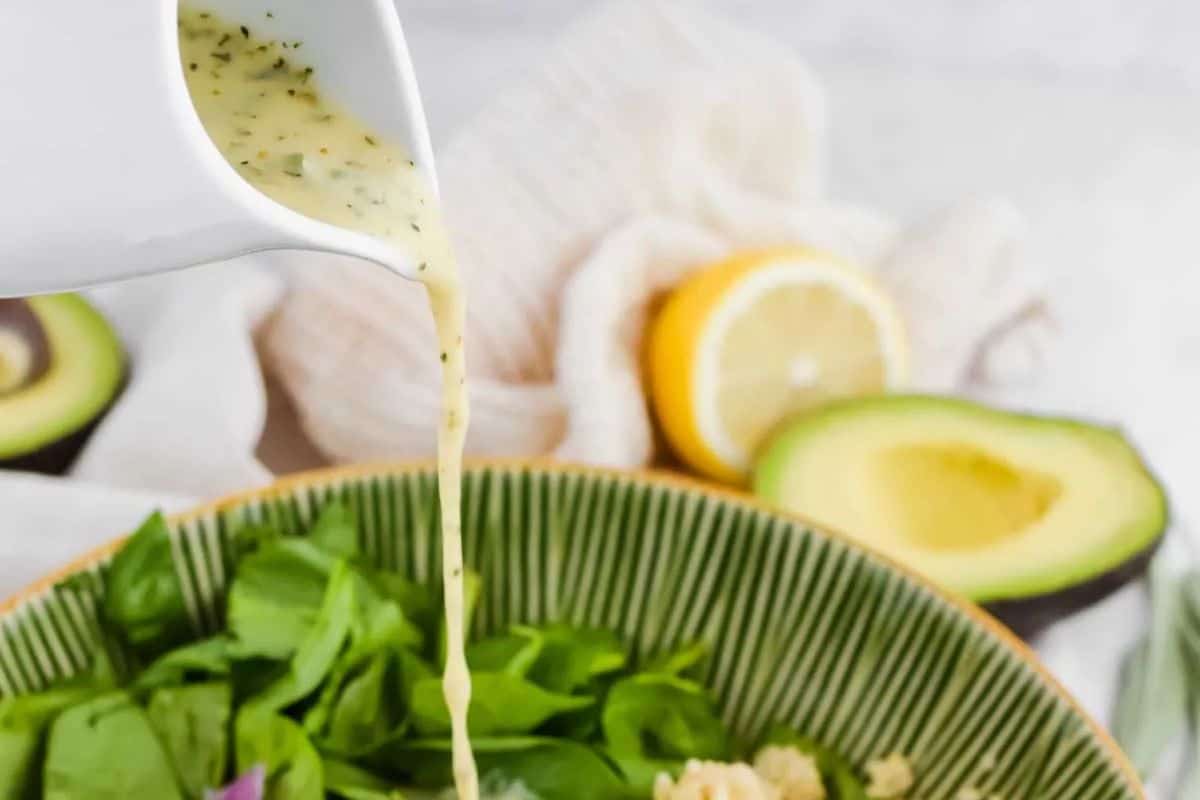 Lemon mustard vinaigrette being poured from a white pitcher into a salad bowl, showing its light yellow color and visible herbs.