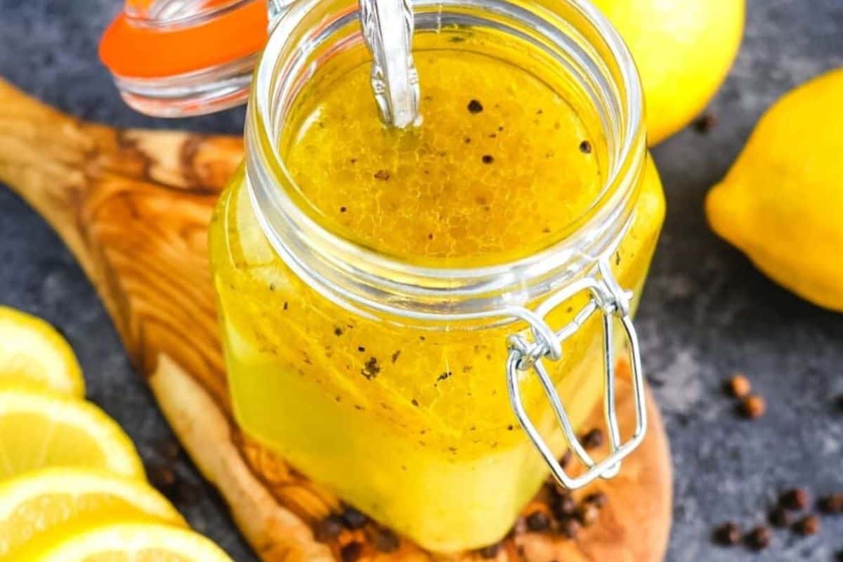 Lemon olive oil dressing in a glass jar with a spoon, surrounded by lemon slices, showing its bright yellow color and visible herbs.