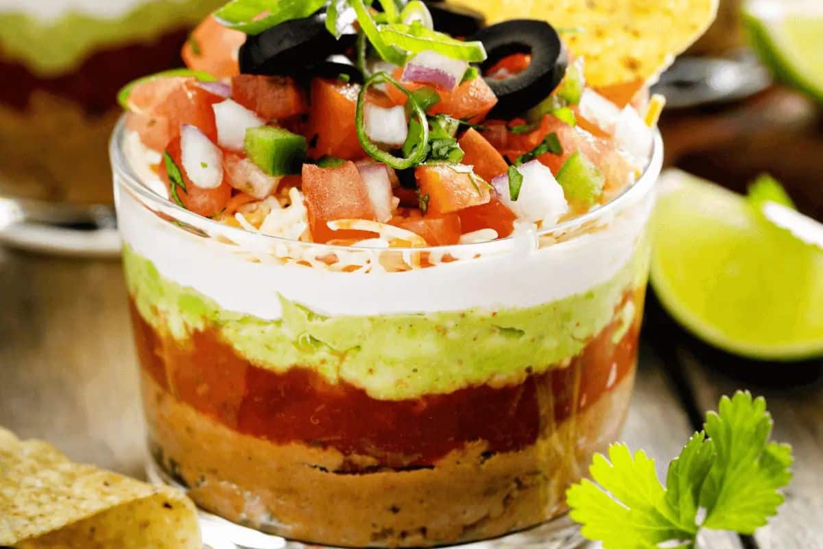 A flavor-packed dip perfect for a Cinco de Mayo party, this layered delight features refried beans, guacamole, sour cream, salsa, shredded cheese, diced tomatoes, green onions, and black olives—all ready to scoop up with crunchy tortilla chips. Lime and cilantro add the final festive touch.