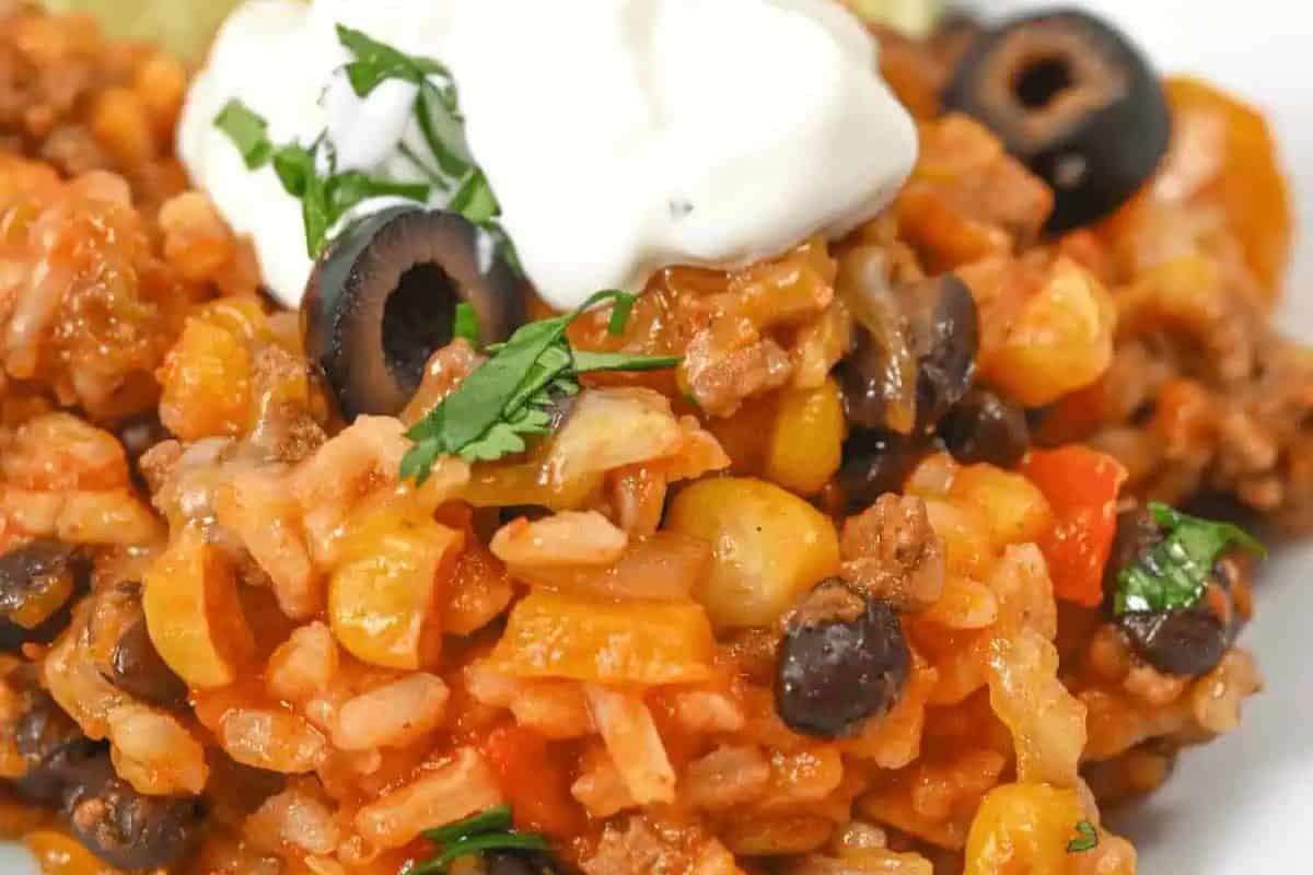 Close-up of a colorful taco casserole reminiscent of vibrant Cinco de Mayo celebrations, featuring ground beef, rice, black beans, corn, diced tomatoes, and black olives. It's topped with sour cream and cilantro for a festive touch.