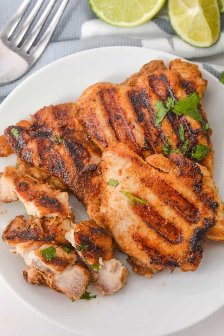 Grilled chicken thighs marinated in a zesty Mexican blend are garnished with fresh herbs on a white plate, complemented by lime slices in the background.