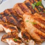 Grilled chicken breast on a white plate, infused with a zesty Mexican Chicken Marinade. Perfectly sliced with grill marks and garnished with green herbs. Juicy slices of lime await in the background, adding a burst of freshness.