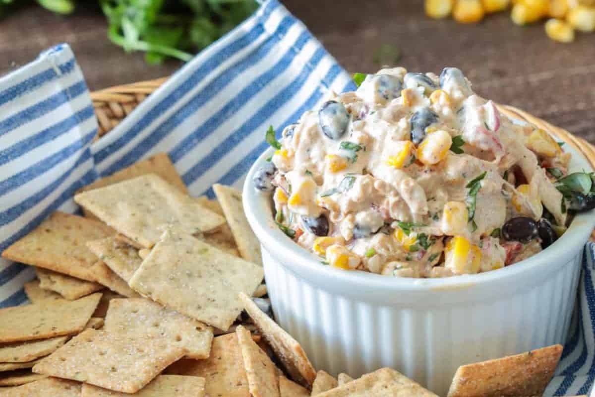 A white ramekin filled with a flavor-packed dip of Mexican chicken salad dip sits on a blue-striped napkin, perfect for Cinco de Mayo festivities. Nearby, wholegrain crackers await in a basket, ready to join the party.