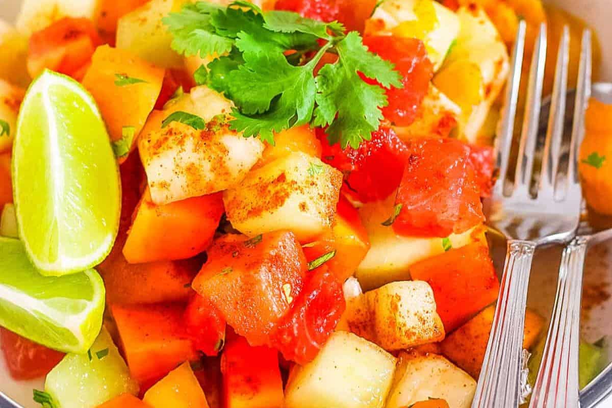 Cubed fruits with chili powder, garnished with cilantro, on a festive plate perfect for Cinco de Mayo. Lime wedges and two forks are placed beside the vibrant fiesta treat.