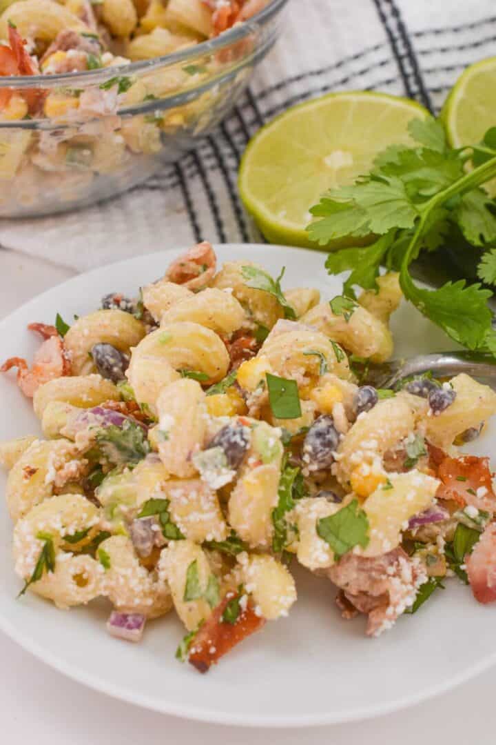 A vibrant Mexican Street Corn Pasta Salad is presented, bursting with beans, tomatoes, and fresh herbs alongside lime halves and cilantro on a checked napkin.