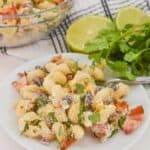 A plate of creamy Mexican Street Corn Pasta Salad with tomatoes, beans, and cheese, garnished with cilantro. A bowl of the same vibrant salad is in the background, accompanied by lime halves and cilantro sprigs.