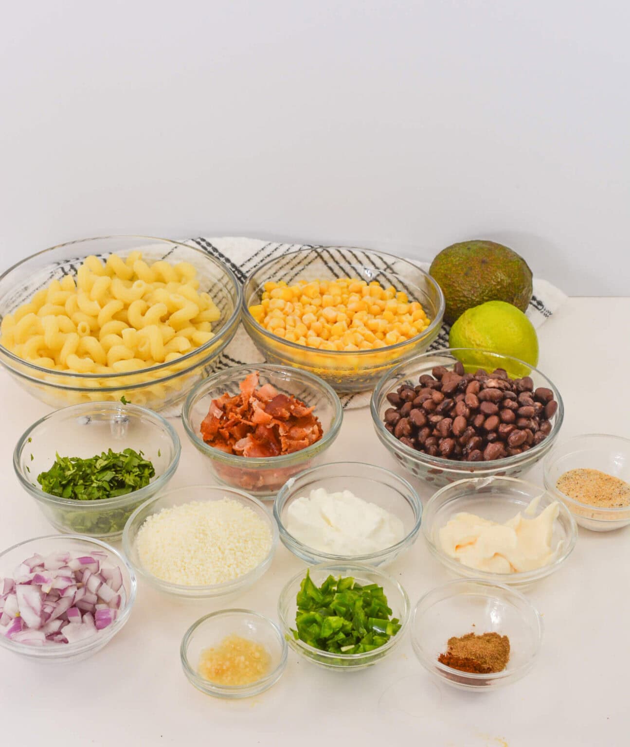 An enticing spread of ingredients in glass bowls featuring pasta, corn, black beans, diced tomatoes, chopped onions, cilantro, and spices on a white surface. Completed with avocado and lime.