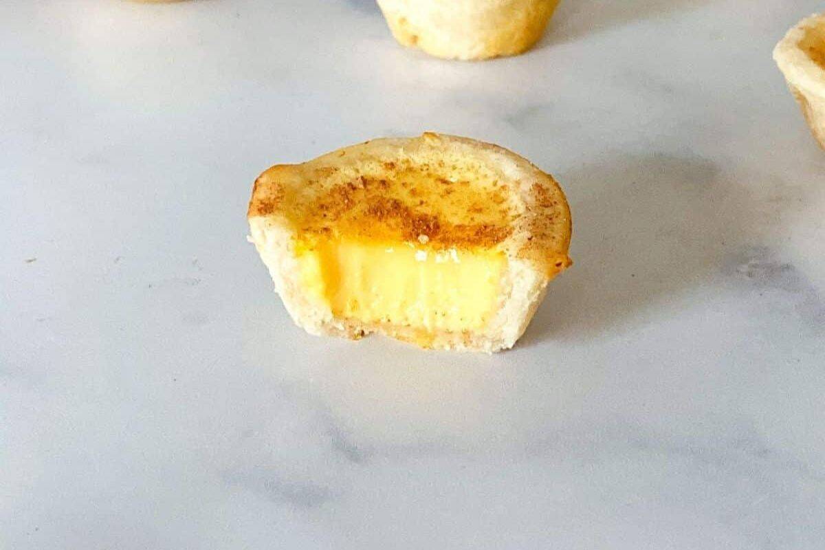 A bitten egg custard tart, reminiscent of adorable recipes, is topped with a sprinkle of cinnamon on a light surface.