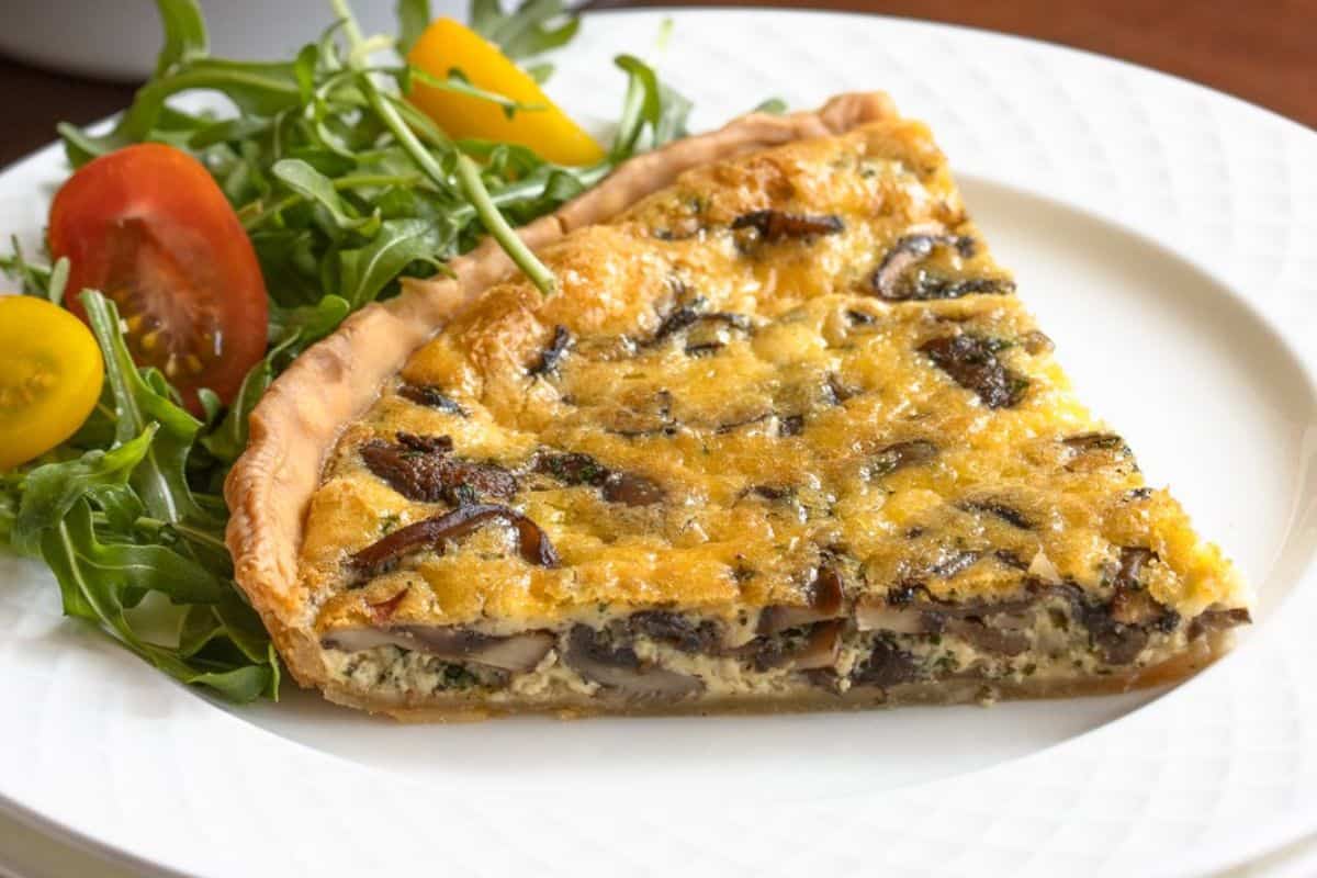 A slice of mushroom quiche sits elegantly on a white plate, paired with arugula, cherry tomatoes, and yellow tomatoes—a perfect addition to your Easter brunch recipes.