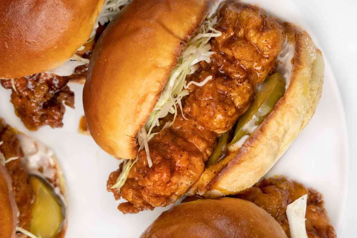 Fried Nashville hot chicken sandwiches with shredded cabbage and pickles on small buns, viewed from above on a white plate.