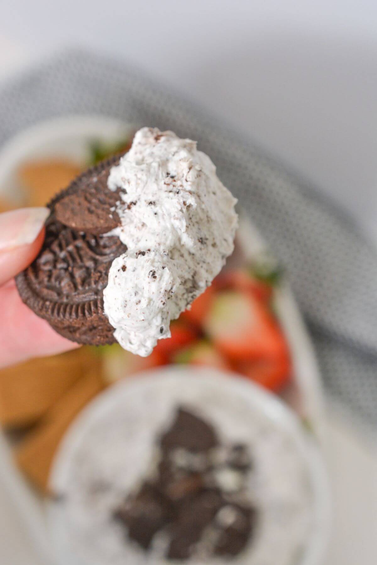 A hand holding an Oreo sandwich cookie dunked in luscious Oreo dip, with a bowl of creamy dip and sliced strawberries artistically blurred in the background.