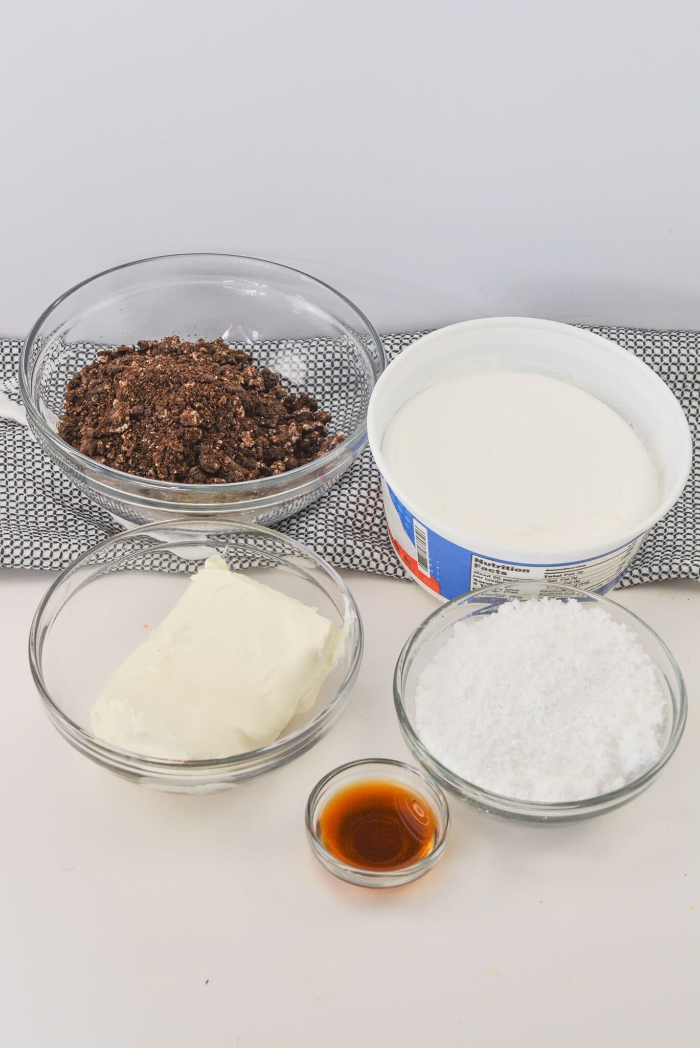 Five glass bowls rest on a white surface: one brimming with crushed chocolate cookies for that perfect Oreo Dip, another with whipped topping, one with smooth cream cheese, powdered sugar in the next, and a small bowl holding fragrant vanilla extract.