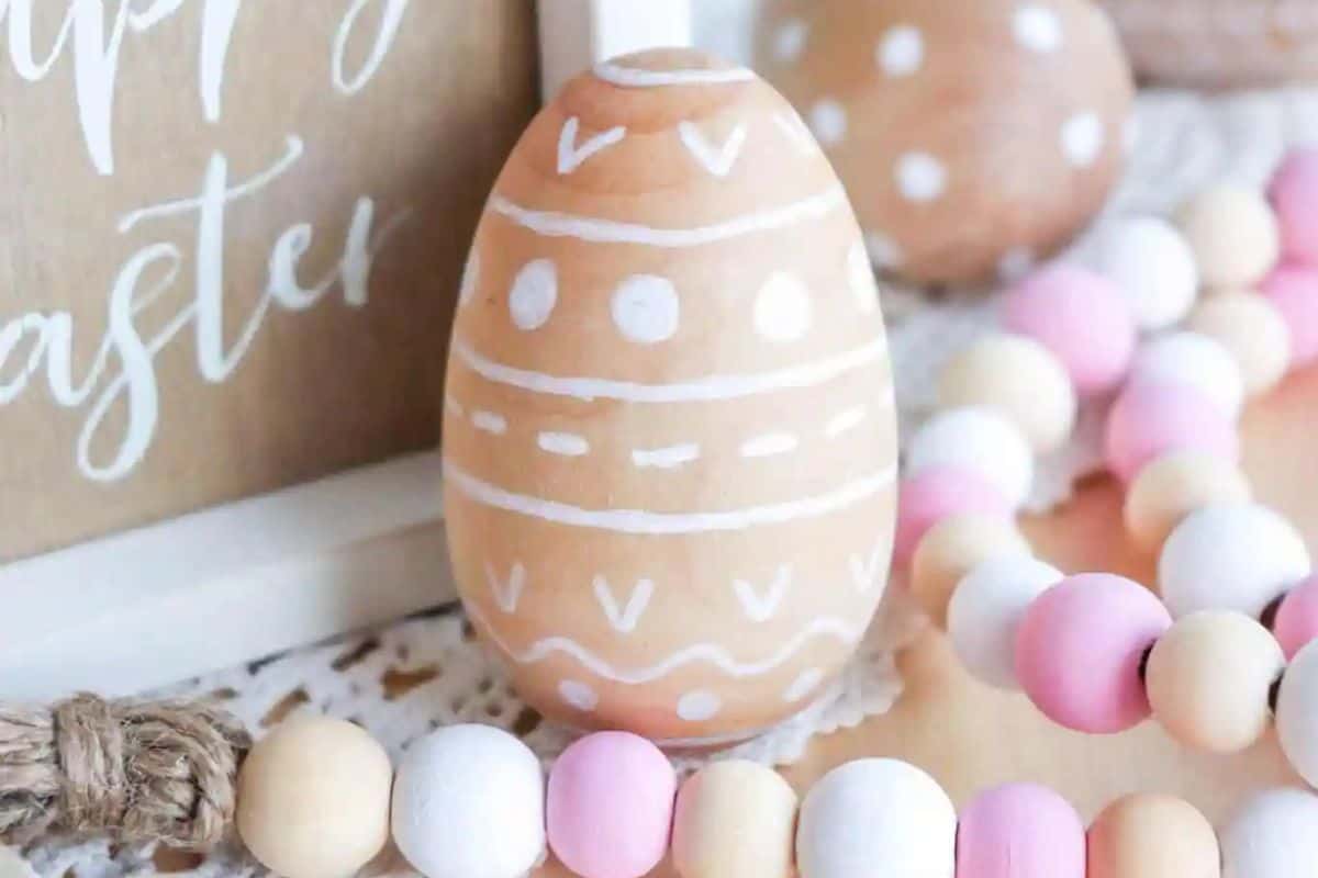 A creatively decorated Easter egg with white patterns is surrounded by pink and white wooden beads. A festive sign peeks through the background, adding to the Easter charm.