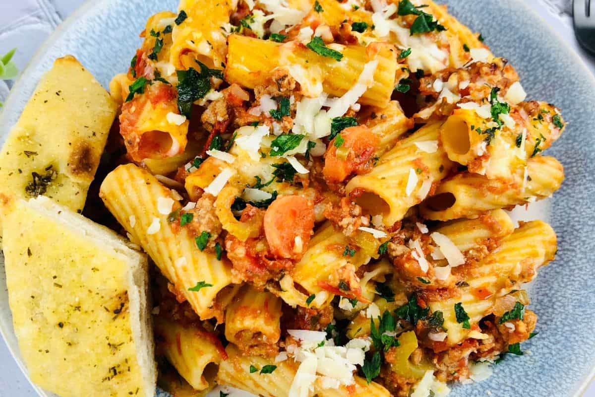This must-try spring plate features rigatoni pasta glazed with tomato sauce and ground meat, sprinkled with fresh herbs and cheese. It's garnished with grated cheese, parsley, and accompanied by savory garlic bread on the side—the ultimate delight in pasta dishes.