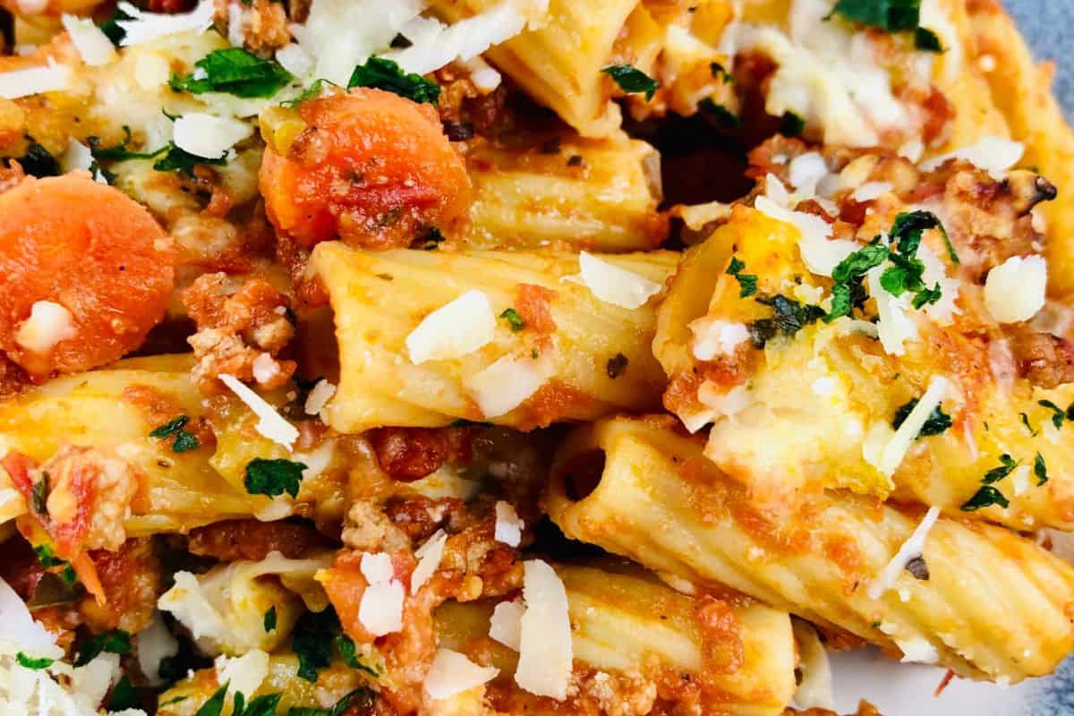A perfect dish for beginners, this plate of rigatoni pasta features ground meat, sliced carrots, sprinkled cheese, and a garnish of chopped herbs.