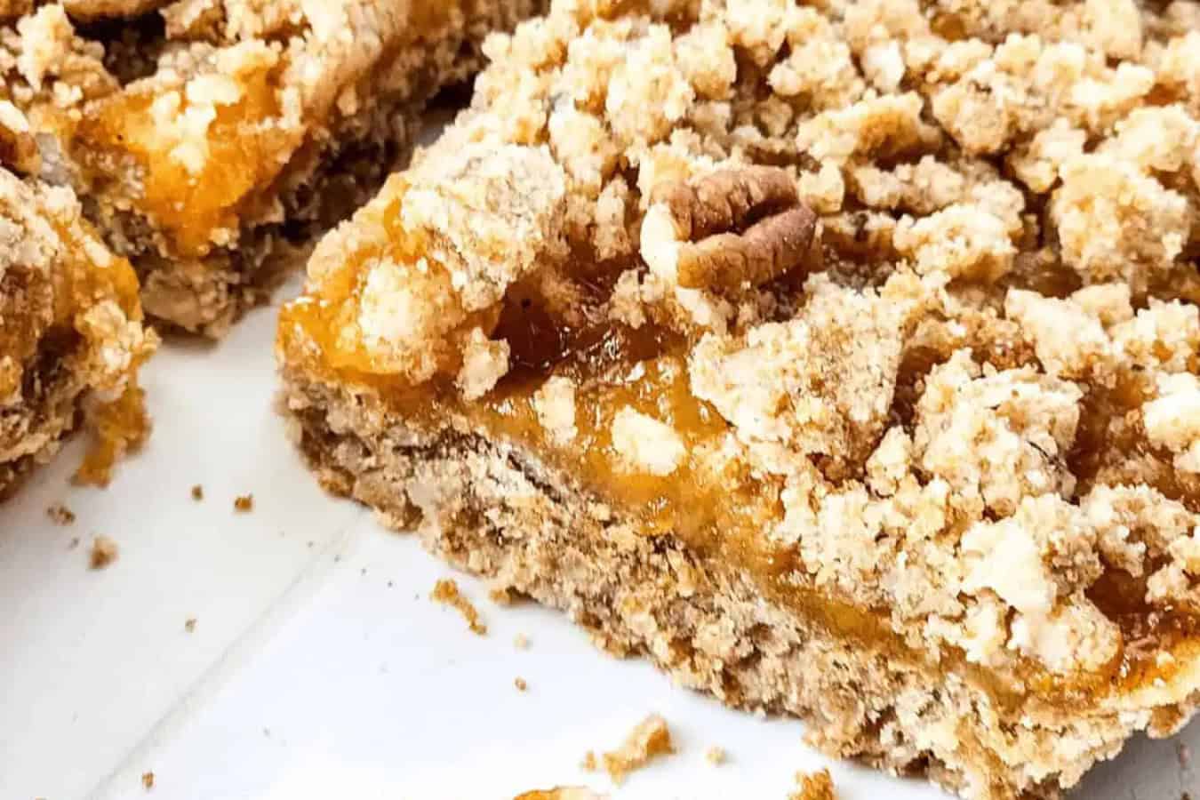 A close-up of a beautiful peach bar with a golden, sticky filling and a crumb topping, revealing layers of crust and topping on a white surface—a true highlight for those who adore tasty desserts.