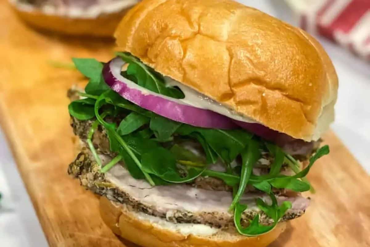 Porchetta sandwich with sliced porchetta, arugula, and red onion on a bun, served on a wooden cutting board.