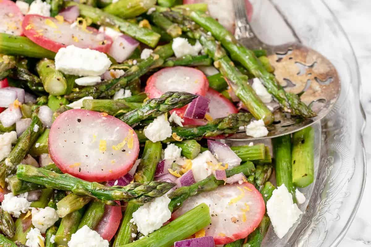 A vibrant spring salad featuring asparagus, sliced radishes, red onion, and crumbled feta cheese is elegantly presented on a clear glass plate with a silver serving utensil—one of the best spring salad recipes to celebrate the season.