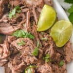 Close-up of Mexican shredded beef garnished with chopped herbs and lime wedges on a white plate.